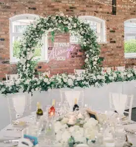 A beautifully decorated wedding reception area with a neon sign reading "It Was Always You." A green floral arch with white flowers frames the sign.