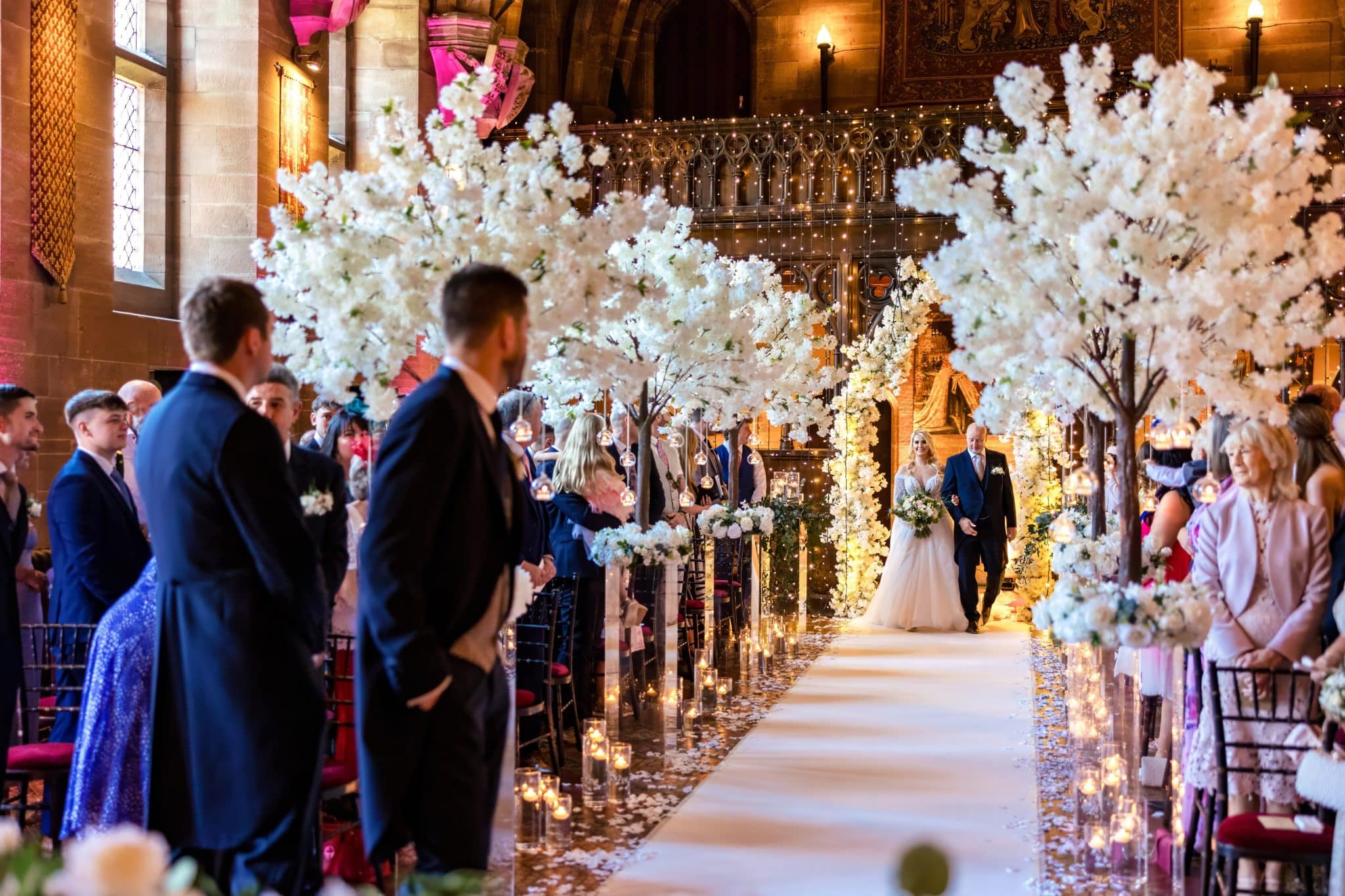 The image shows Lauren walking down the aisle with a man, possibly her father, amid elegant white flower arrangements and candles.