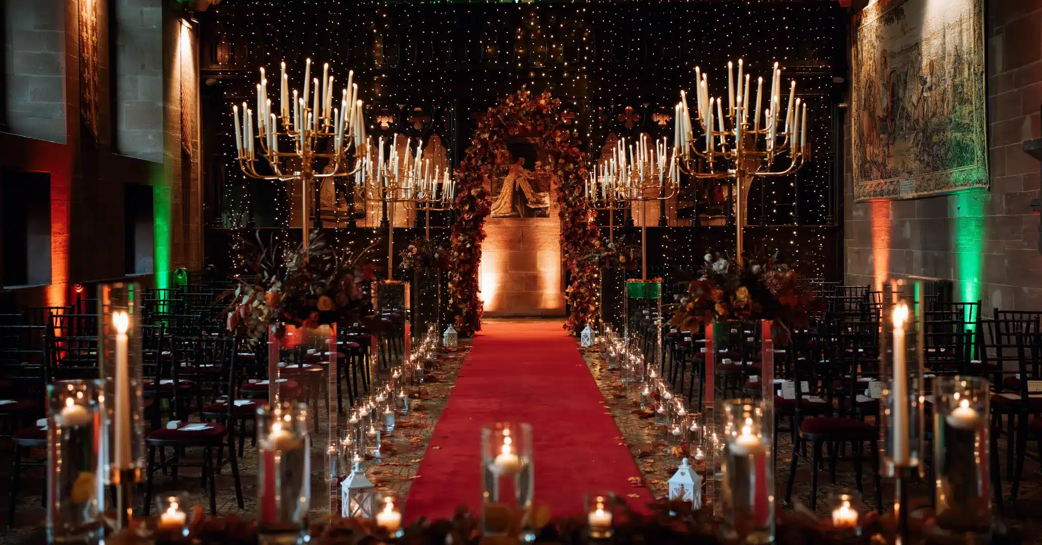 A beautifully styled wedding ceremony setup at Peckforton Castle, featuring warm autumnal tones, elegant gold candelabras, and a romantic red aisle with ambient lighting.